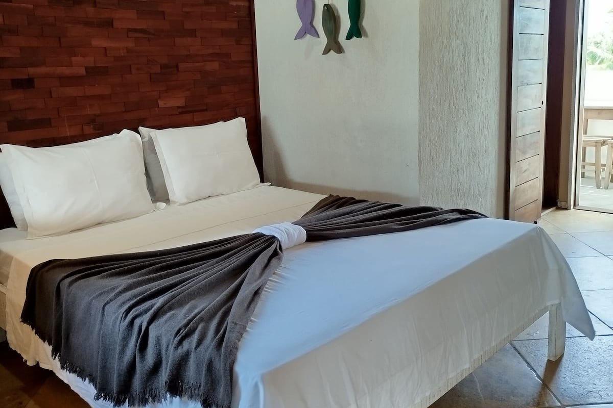 Uma cama com roupa de cama branca e um cobertor cinza escuro, teto de madeira, parede de tijolos e um enfeite de peixe fixado na parede.