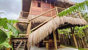 A thatched-roof bungalow with a wooden staircase leading to a balcony, surrounded by lush greenery. - Caravelas Praia (Amontada)
