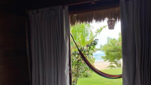 A bed with a brown blanket and white towels, a hammock, and a view of a beach and greenery through a window with curtains. - Caravelas Praia (Amontada)