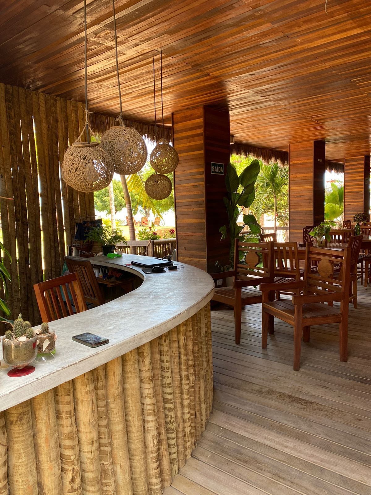 A wooden interior with a curved counter, wicker hanging lights, and a view of greenery outside.
