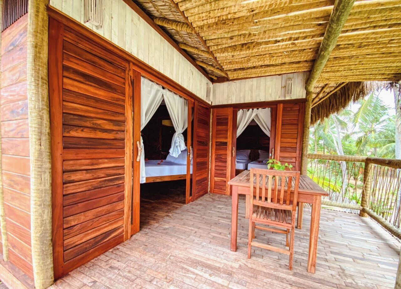 A wooden cabin with a thatched roof, a wooden table and chair, and two open beds with white curtains.