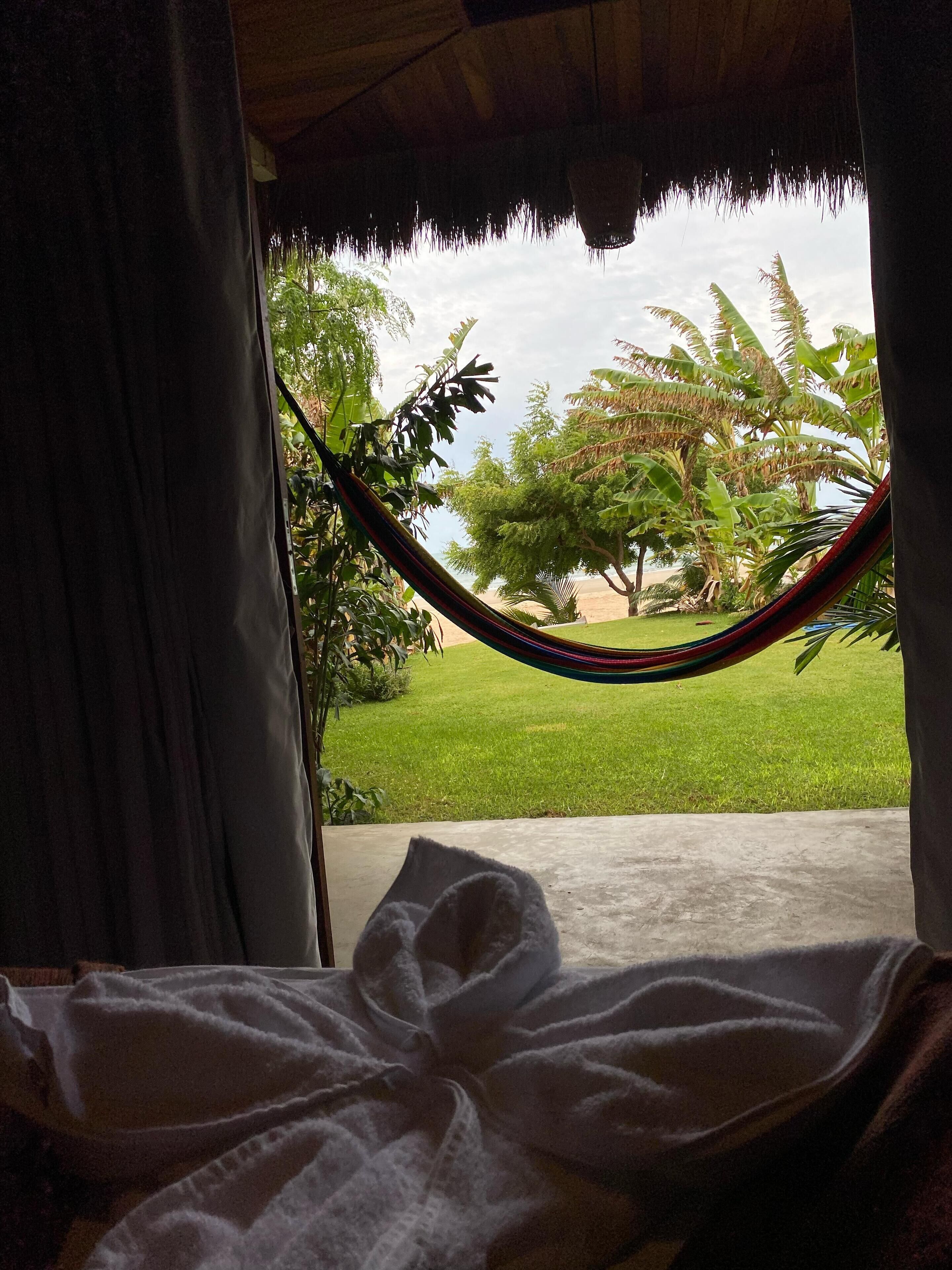 A room with a view of a grassy area and trees, a hammock, and a bed with a white blanket.