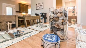 A dining table with a glass vase filled with seashells, a 'BEACH PLEASE' placemat, and a coffee maker in the background.