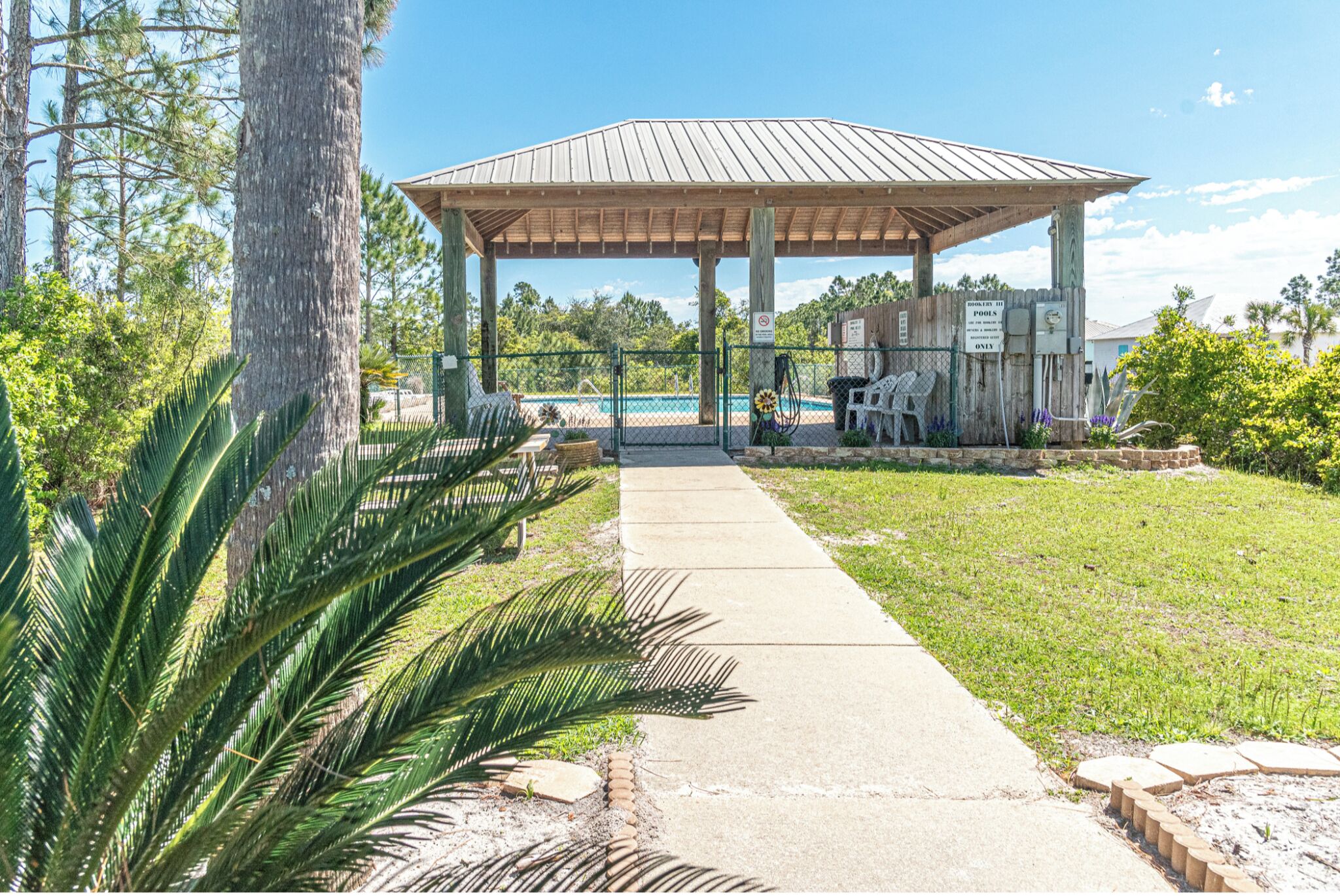 A gazebo with a concrete pathway leading to a pool area, surrounded by greenery and a wooden fence.