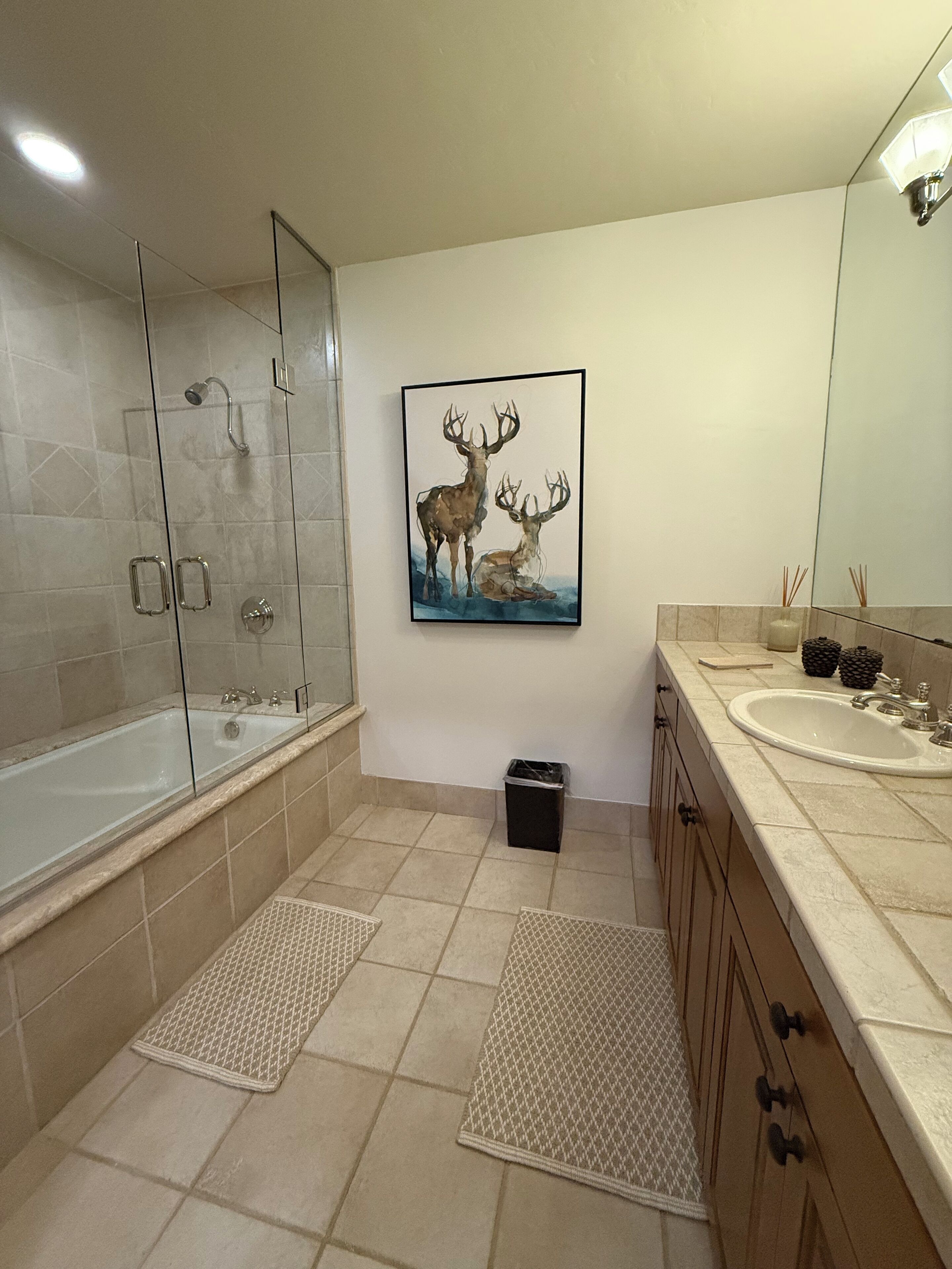 A modern bathroom with a glass-enclosed shower, a large mirror, a sink with a countertop, and a wall-mounted artwork of deer.