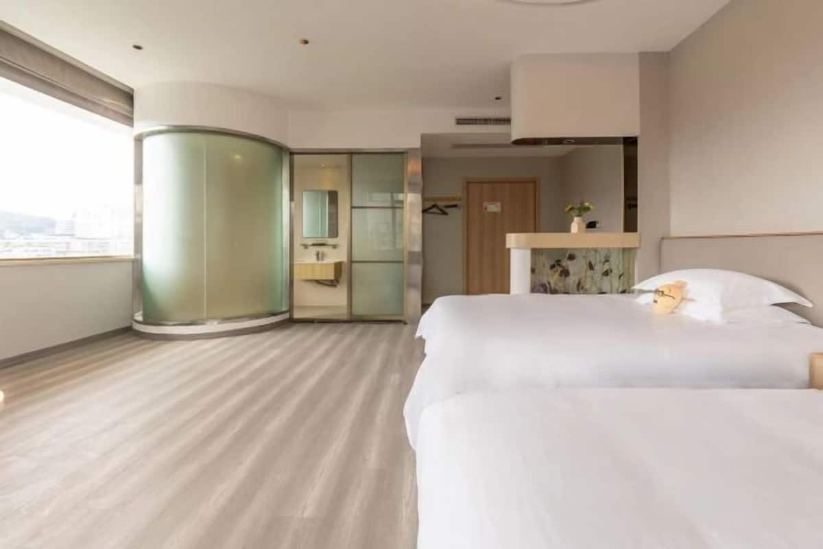 Habitación de hotel con dos camas, un separador de vidrio curvo, una puerta de madera y vista a la ciudad.