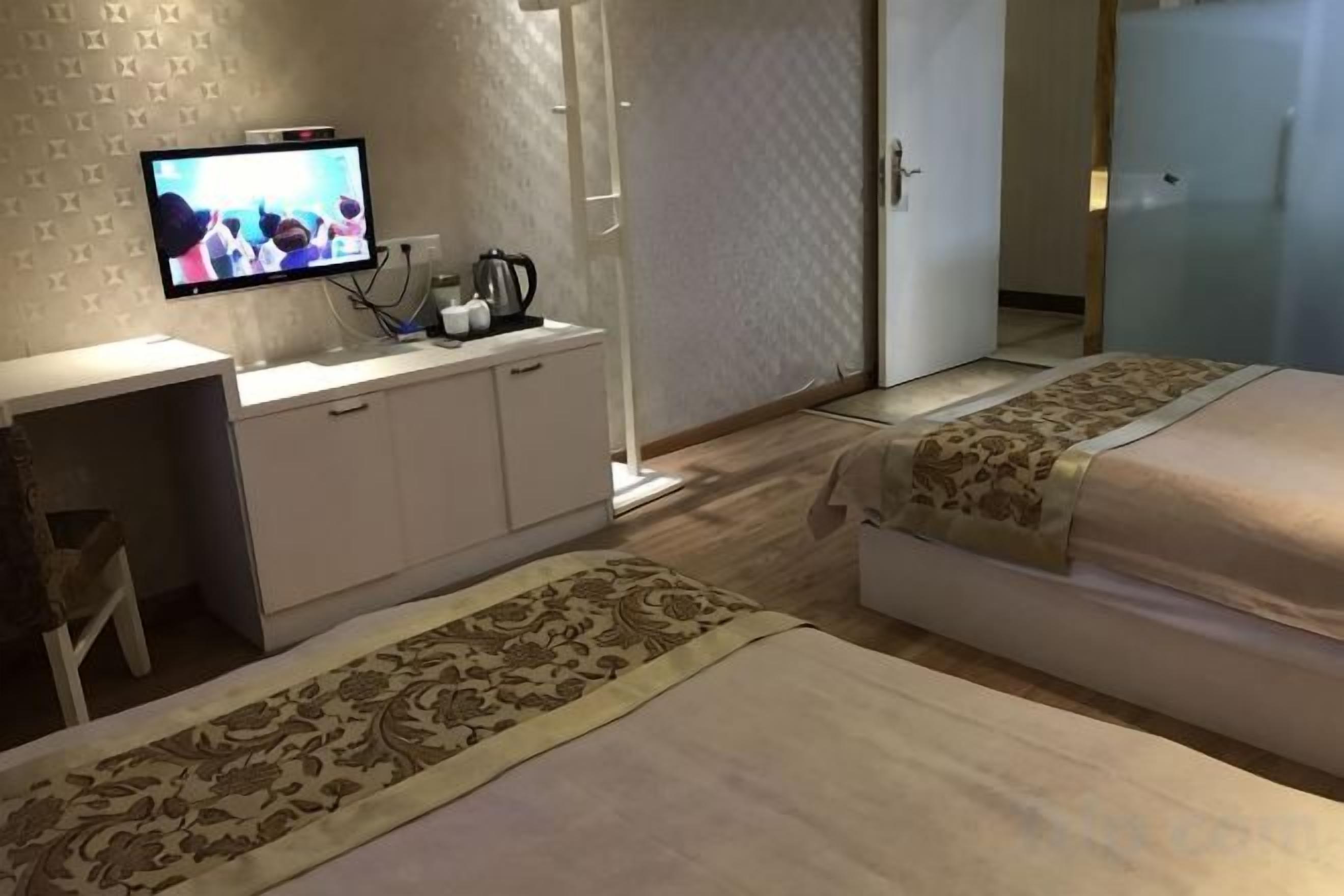 Ein Hotelzimmer mit Fernseher auf einem Schrank, einem Schreibtisch mit Stuhl, zwei Betten mit gemusterten Tagesdecken und einem Teppich mit Muster.