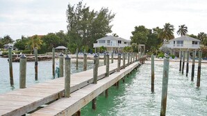 A wooden pier extending into calm water, supported by wooden posts, with a row of beachside houses in the background.