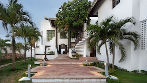 A modern building with a white exterior, a central staircase leading to a door, and a surrounding garden with palm trees and a paved walkway. - Villa de Lujo en Primera Línea de Playa (San Luis)