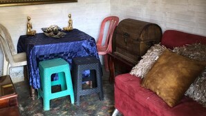 Een kamer met een rode bank, een houten kist, een tafel met een blauwe tafelkleed, twee krukken en een schilderij aan de muur.
