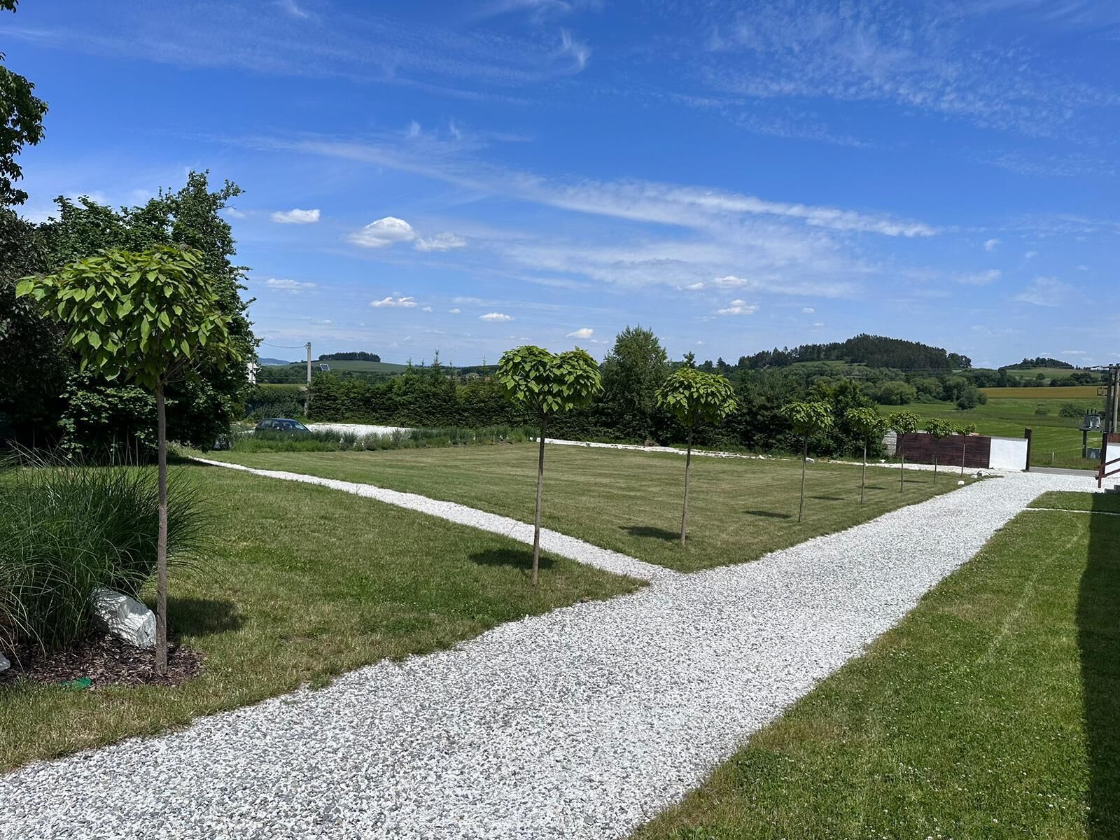 A grassy area with a gravel path, trees, and a distant view of hills.