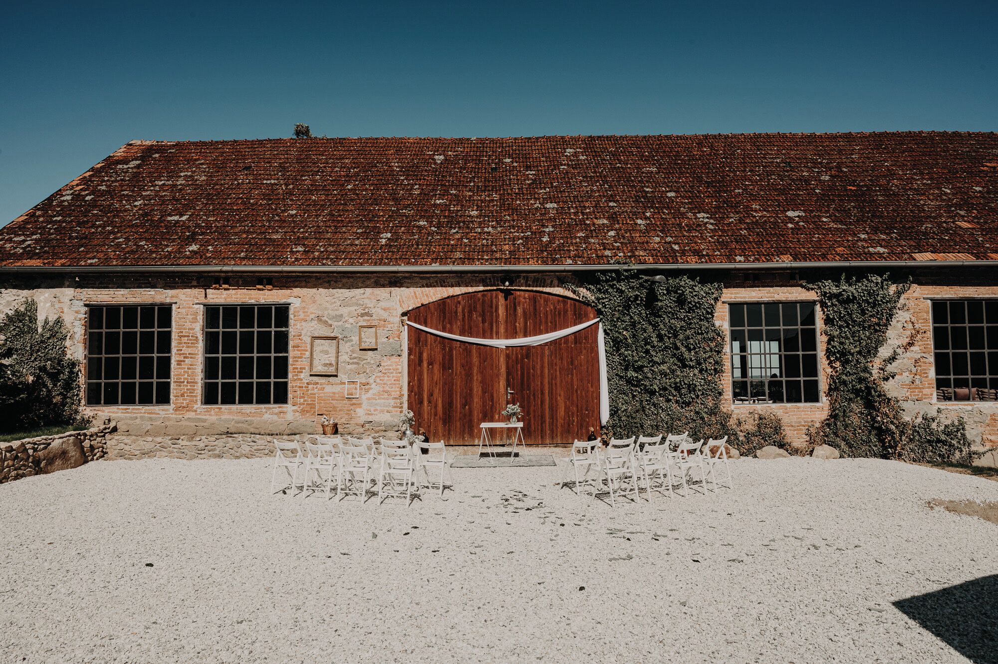 A rustic building with a tiled roof, a large wooden door, and a gravel courtyard with white chairs.