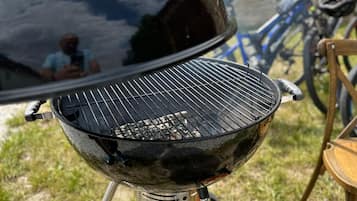 A portable grill with a lid, set on a grassy area with a bicycle and a chair nearby.