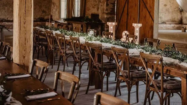 A long dining table set with chairs, candles, and greenery in a rustic room with exposed brick walls and wooden beams. - Chateau Kravín (Kurimany)