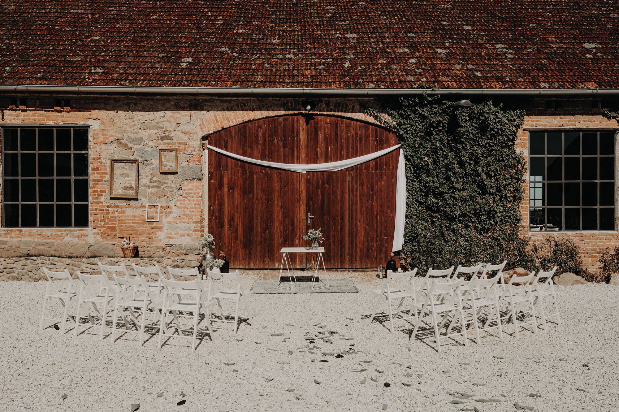 Montaje para una boda al aire libre con un granero rústico de fondo, sillas blancas dispuestas en filas, una pequeña mesa con un florero y una puerta de madera.