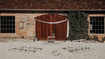 Montaje para una boda al aire libre con un granero rústico de fondo, sillas blancas dispuestas en filas, una pequeña mesa con un florero y una puerta de madera.