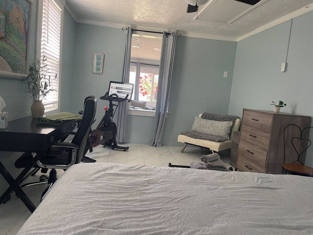 A bedroom with a bed, a desk, an exercise bike, a chair, a dresser, and a window with curtains.
