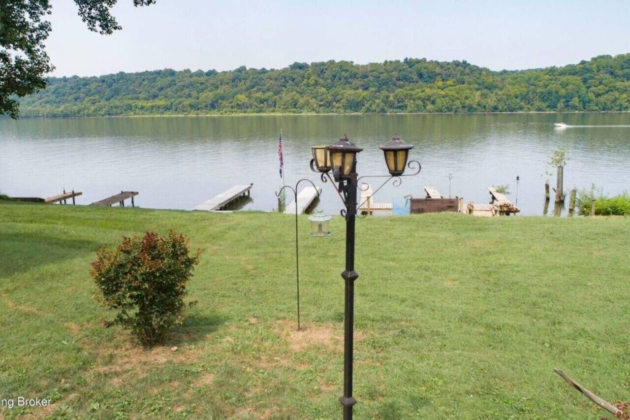 A grassy area with a lamp post and a dock extending into a lake, surrounded by trees.
