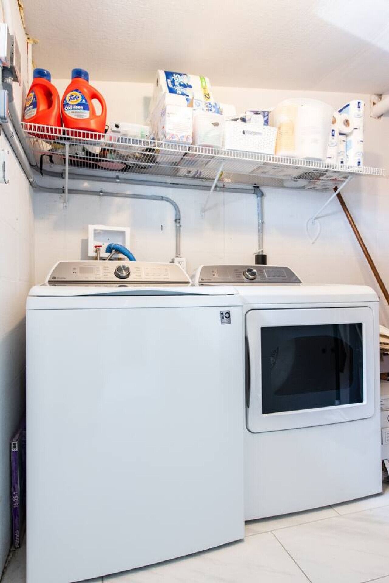 Un espace buanderie comprenant un lave-linge et un sèche-linge à chargement frontal, une étagère avec du détergent et des essuie-tout, et un chauffe-eau.