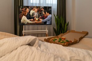 Una habitación con una cama, un televisor mostrando un grupo de personas y una caja de pizza sobre la cama.