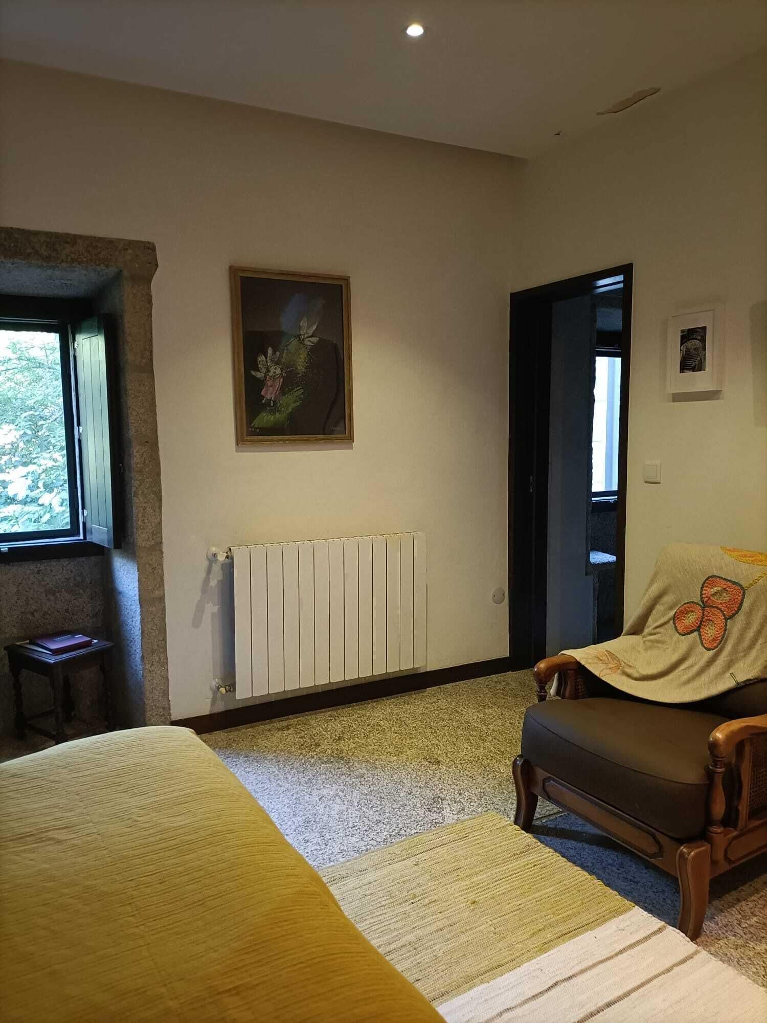 Un dormitorio con cama, una silla, un radiador, una ventana y un cuadro en la pared.