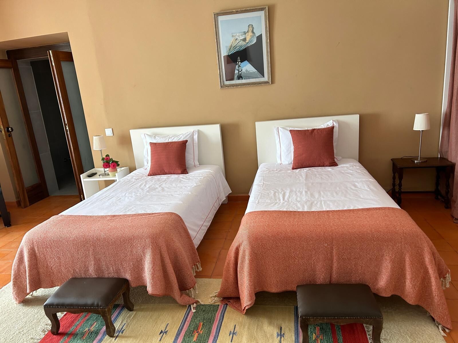 Quarto de hotel com duas camas, uma mesinha com abajur, um banco e um quadro na parede.