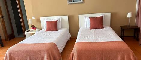 Quarto de hotel com duas camas, uma mesinha com abajur, um banco e um quadro na parede.