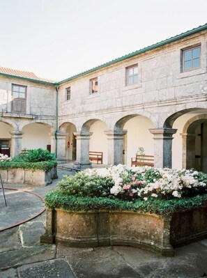 Un patio con edificios de piedra, pasillos abovedados y macizos de flores.