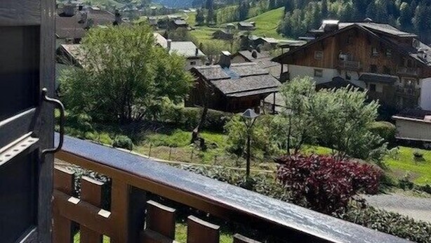 A balcony with a view of a mountain village, featuring traditional houses and snow-capped peaks.