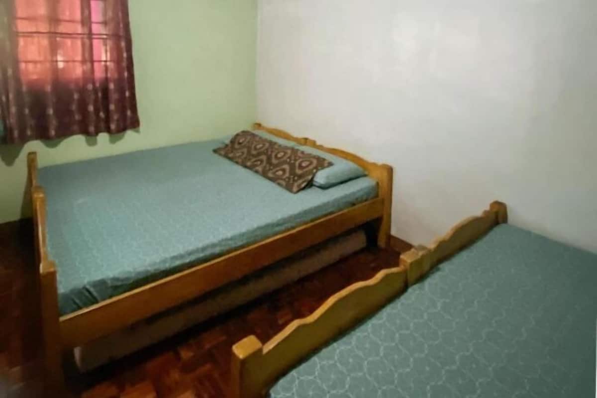 Ein Einzelbett mit Holzrahmen, ein gemustertes Kopfkissen, ein kleiner Nachttisch und ein Fenster mit Vorhang.
