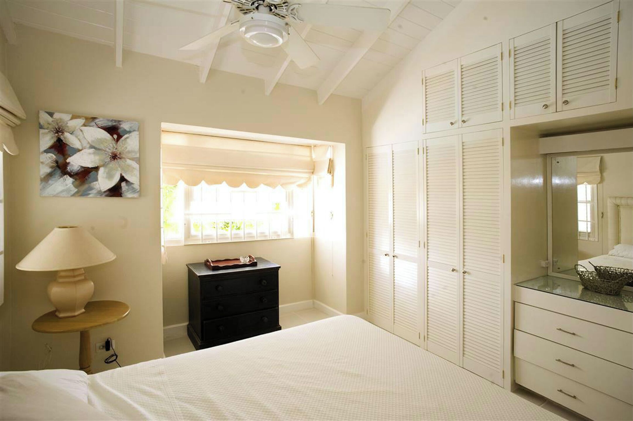 A bedroom with a bed, a dresser, a nightstand, a lamp, a window with blinds, a ceiling fan, and a wardrobe.