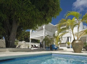 A pool with lounge chairs, a large tree, and a building with a balcony. - Mango Court #1 - by Zenbreak (Holetown)