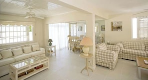 A spacious living room with a white sofa, a checkered sofa, a dining area with a table and chairs, and a ceiling fan. - Mango Court #1 - by Zenbreak (Holetown)