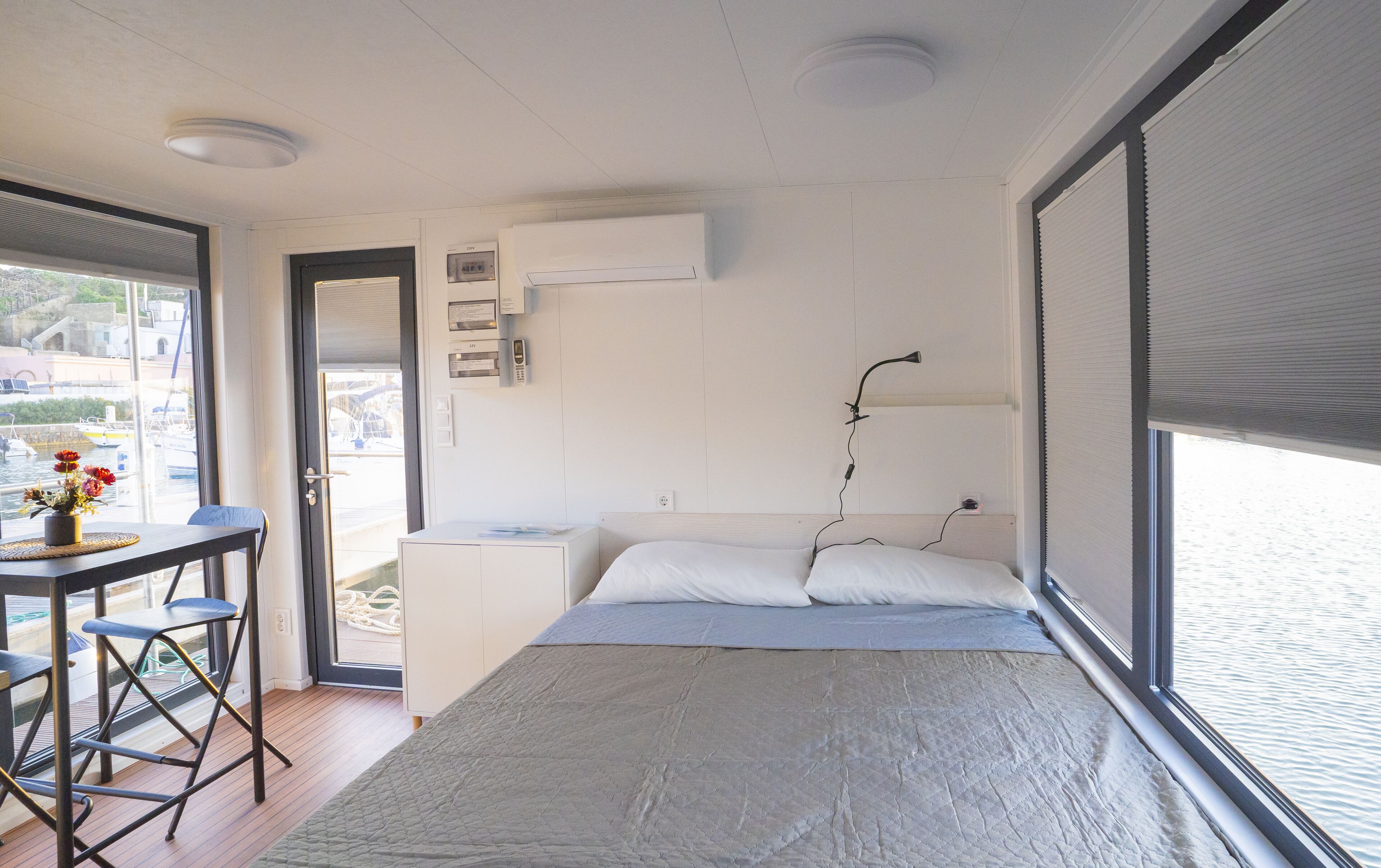 A bedroom with a bed, a desk, and a chair. There is a window with blinds, a door, and a view of boats outside.