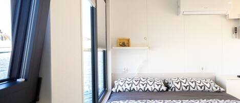 Une chambre d’hôtel avec un grand lit, une fenêtre avec des stores, un climatiseur fixé au mur et une petite étagère sur laquelle se trouve un cadre photo.