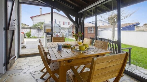 A covered patio with a wooden table and chairs, a glass door leading to a backyard, and a view of neighboring houses. - Maison Confort 3 Chambres Piscine- 6 Couchages, Tarnos Limite Pays Basques (Tarnos)