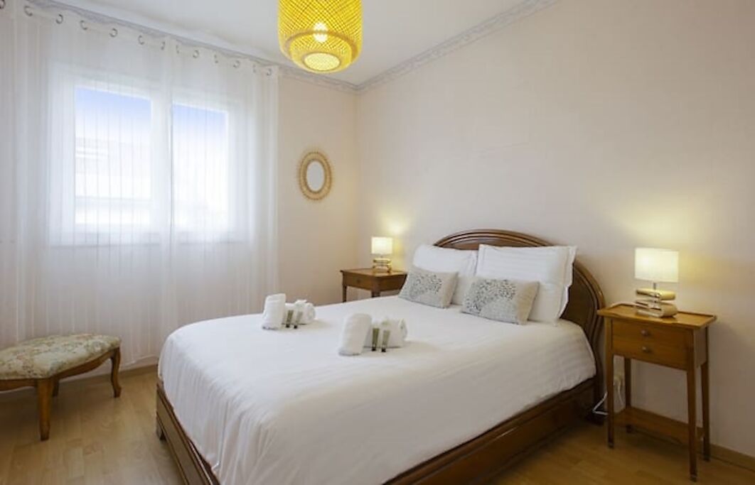 Ein Hotelzimmer mit einem großen Bett, zwei Nachttischen inklusive Lampen, einer kleinen Bank und einem Fenster mit durchscheinenden Vorhängen.