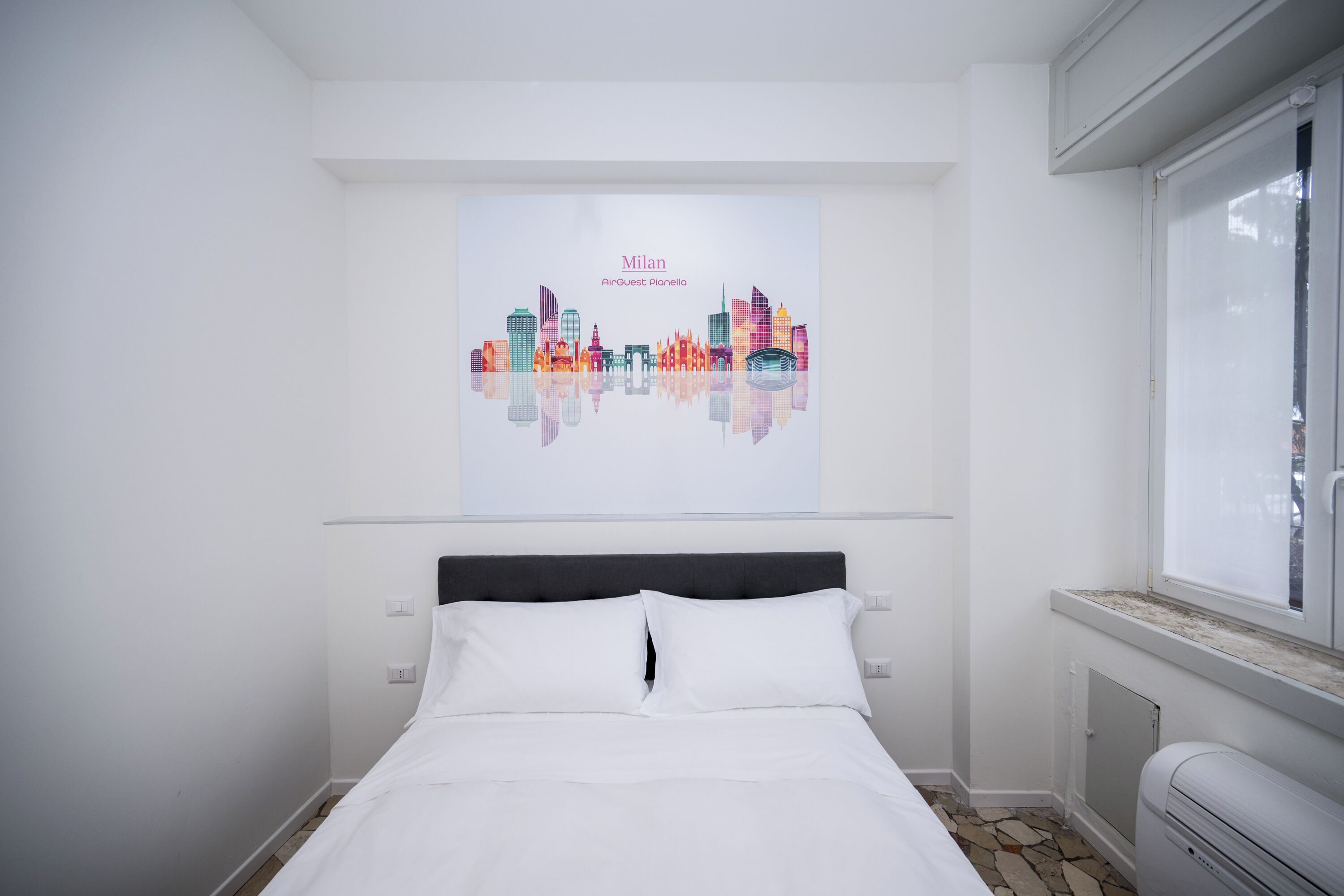 Une chambre d’hôtel avec un lit, une fenêtre et une affiche représentant un paysage urbain accrochée au mur.