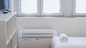 Habitación de hotel con una cama, aire acondicionado blanco, televisor sobre un soporte y una ventana con persianas.
