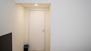 Un pasillo estrecho con una puerta cerrada, un bote de basura y una lámpara en el techo.