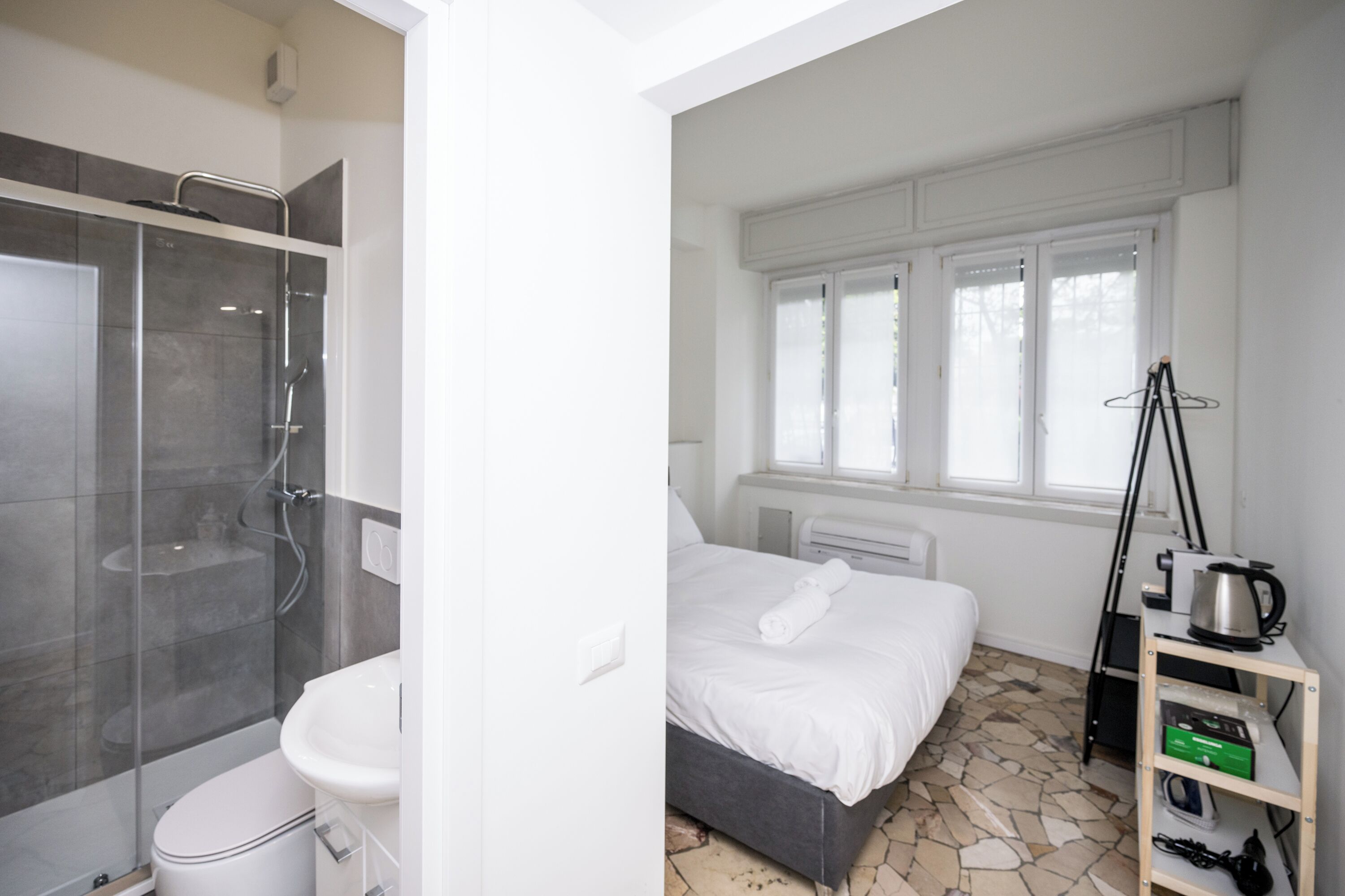 Une salle de bain moderne avec une douche à parois de verre, des toilettes blanches et un lavabo. À côté de la salle de bain se trouve une chambre avec un lit, une fenêtre et une petite étagère sur laquelle sont posés une bouilloire et une boîte verte.