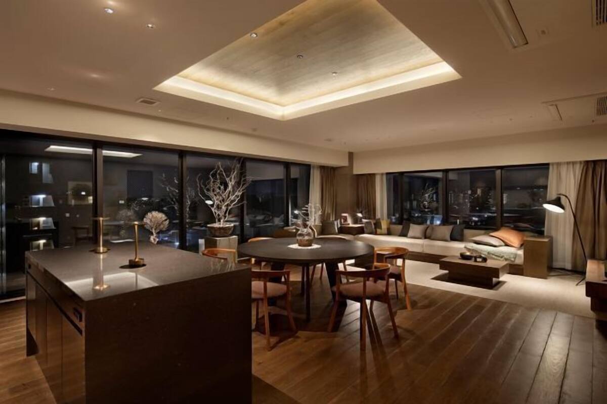 A modern living room with a dining area, large windows, and a high ceiling.