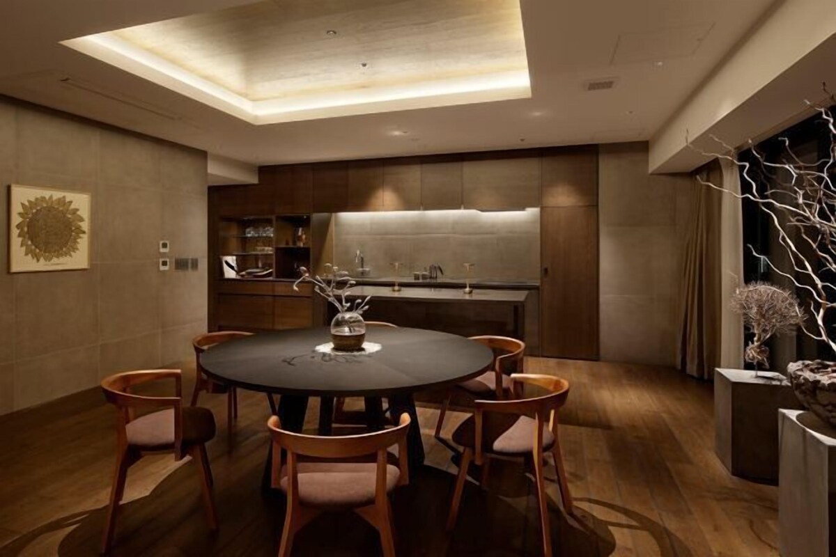 A modern dining area with a round table, six chairs, a ceiling light, and a kitchen in the background.