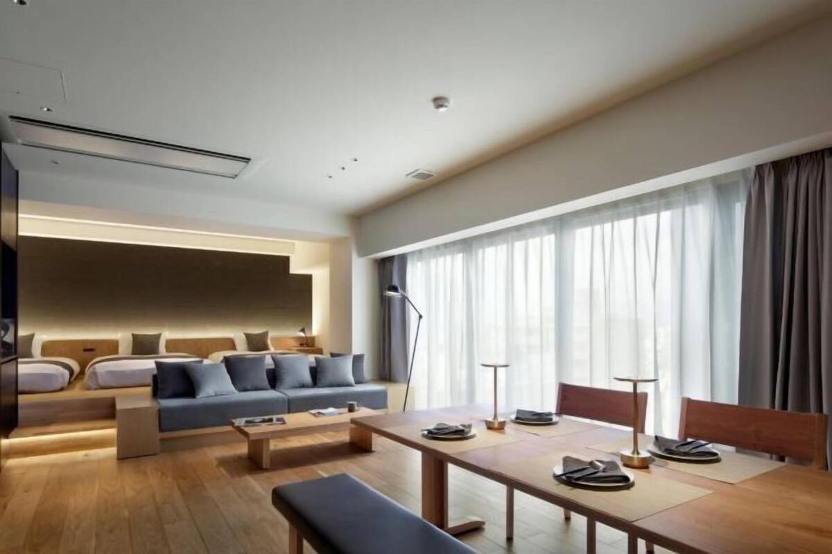A modern hotel room with a sofa, a dining table, and a large window with curtains.