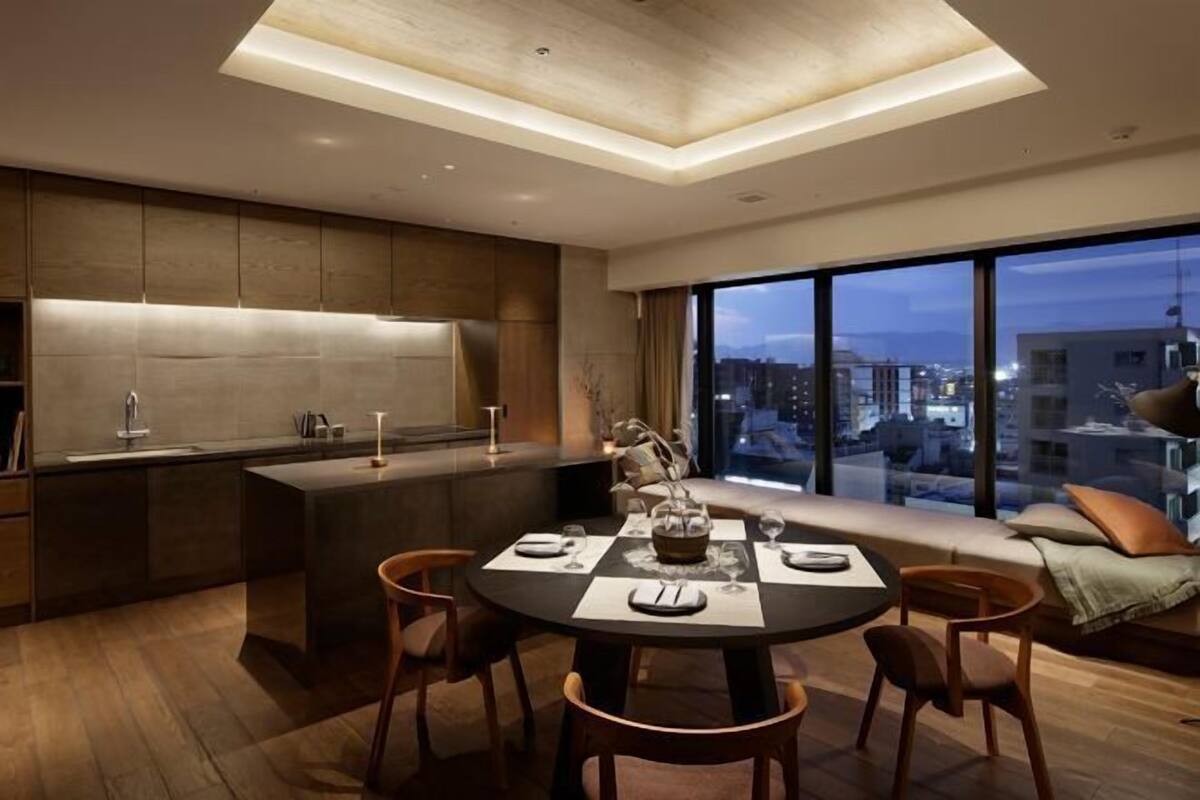 A modern kitchen with a dining area, large windows with a city view, and a built-in bench with cushions.
