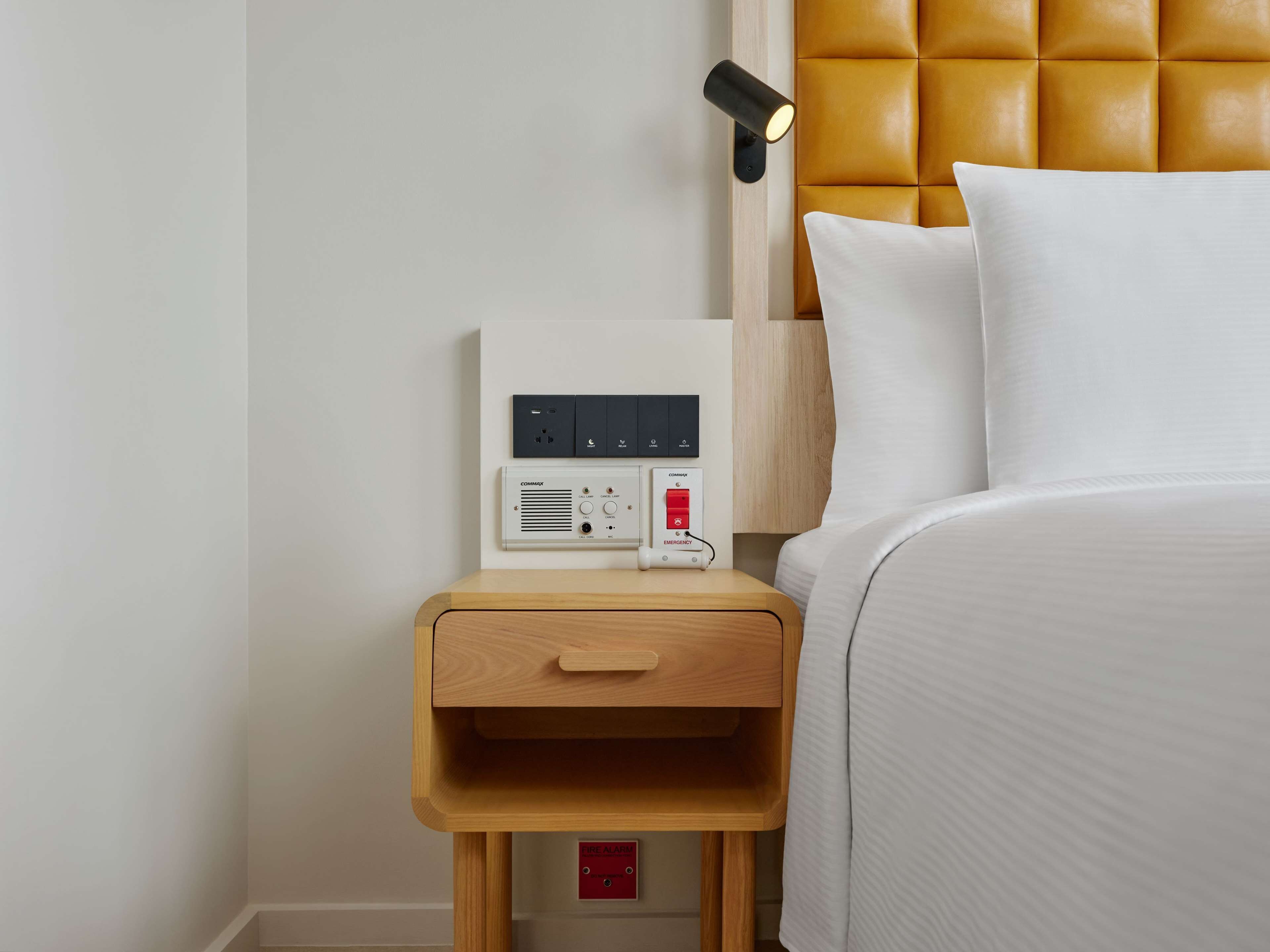 Een hotelkamer met een bed, een nachtkastje met bedieningspaneel, een wandlamp en een rookmelder.