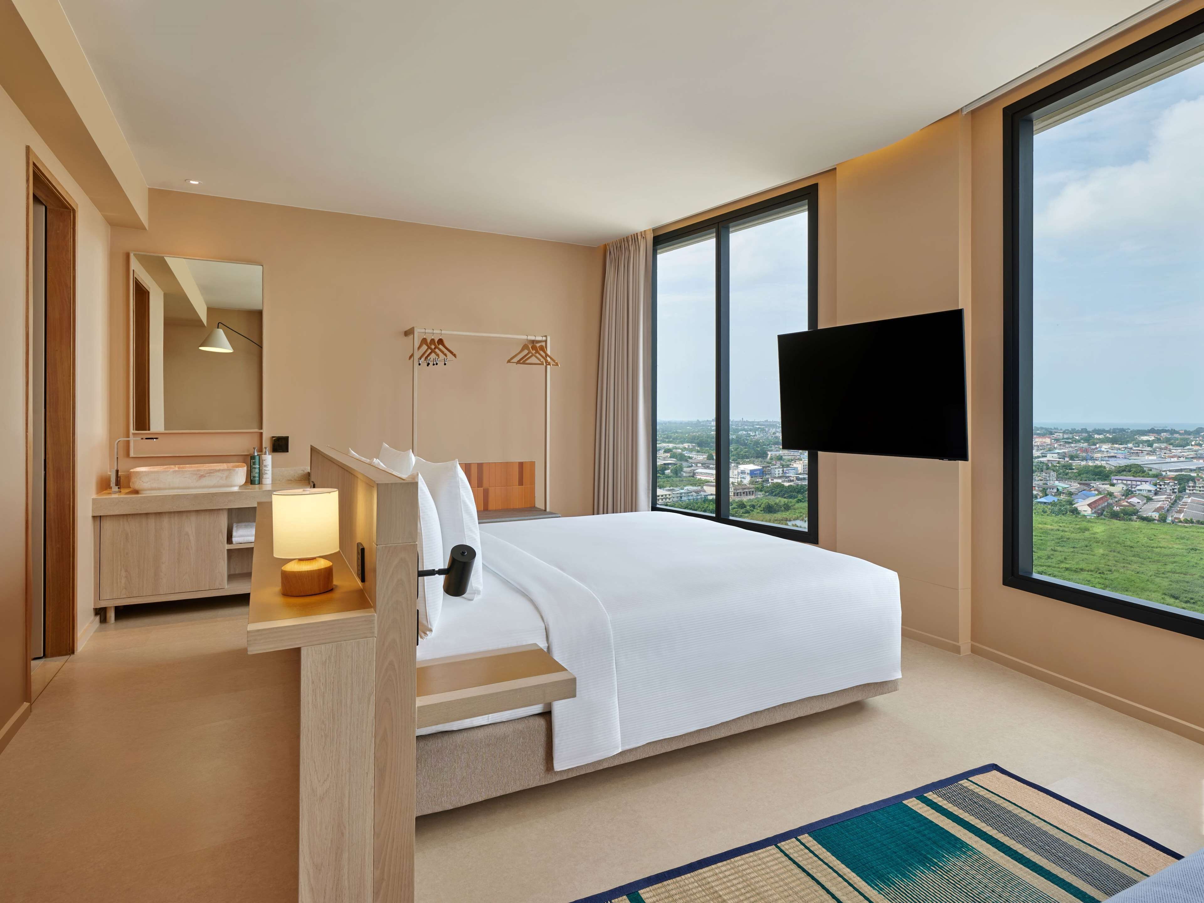 A hotel room with a large bed, a bedside table with a lamp, a mirror, a TV mounted on the wall, and a view of the city through large windows.