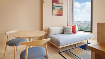 Ein modernes Hotelzimmer mit rundem Tisch, einer Couch mit Kissen, einem großen Fenster mit Blick auf die Stadt und einer Pendelleuchte.