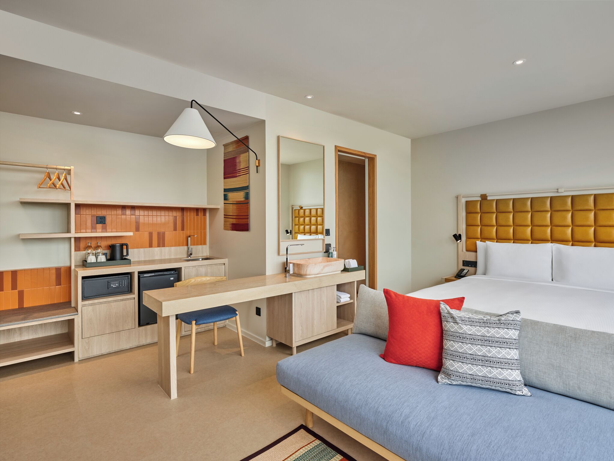 Una habitación de hotel moderna con kitchenette, una cama con cabecera tapizada, un escritorio con silla y un espejo grande.