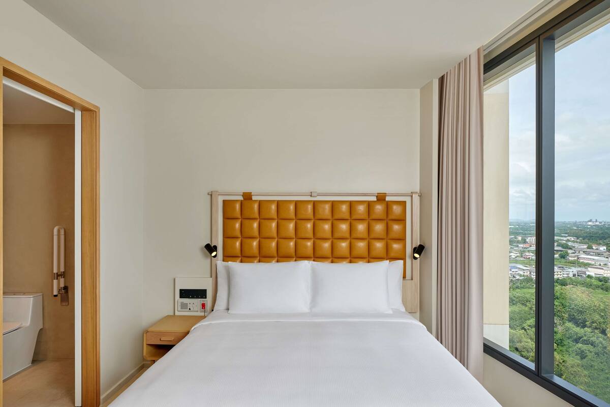 Una habitación de hotel con una cama grande, cabecera de madera, una mesita de noche y un ventanal con vista a la ciudad.